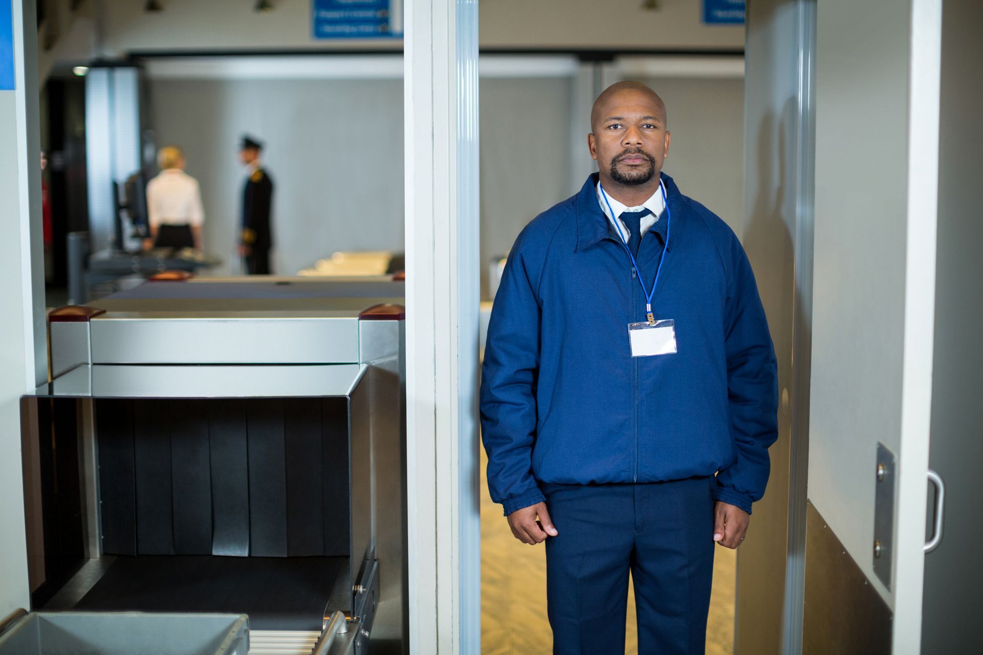 Airport security guard stands in doorway, metal detector visible. Uniformed, wearing ID badge.