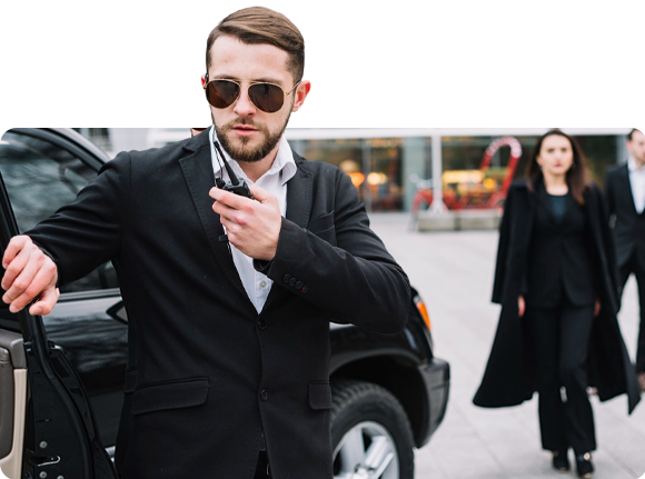 A man in a suit, sunglasses, and a radio by a car, with two people in black behind him.