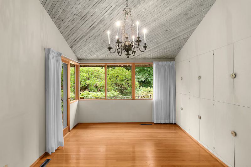 Empty room with wood floors, white walls, angled, wooden-planked ceiling. Large window and built-in white cabinets.