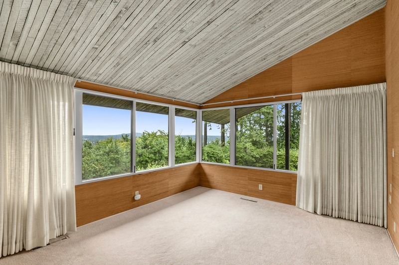 Empty room with large windows, white curtains, wood-paneled walls, and a sloped, textured white ceiling.