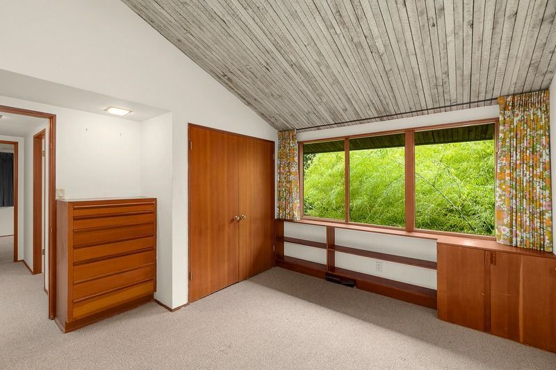 Bedroom with sloped, wood-paneled ceiling, large window with greenery view, light-colored carpet, and wooden cabinetry.
