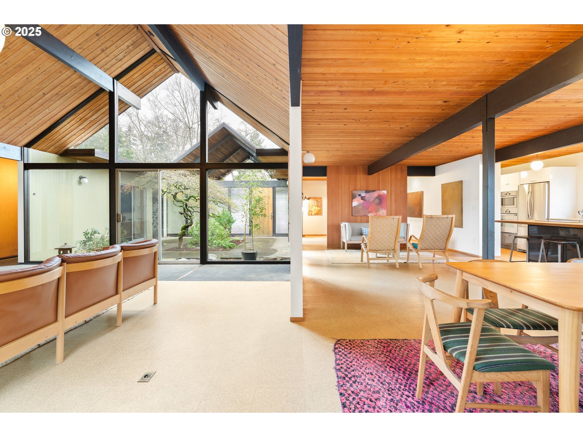 Mid-century modern home interior with floor-to-ceiling windows, wooden beams, and a view of a courtyard.