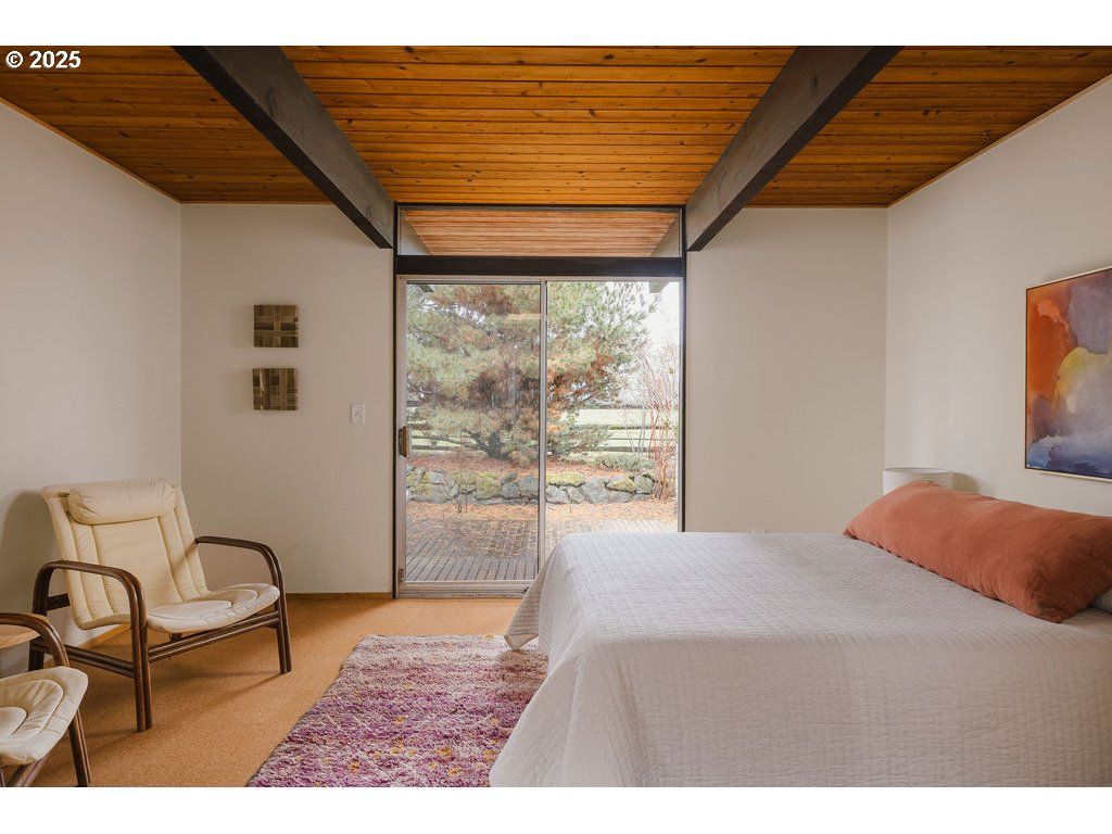 Bedroom with wood ceiling, glass doors, bed, chairs, and colorful rug.