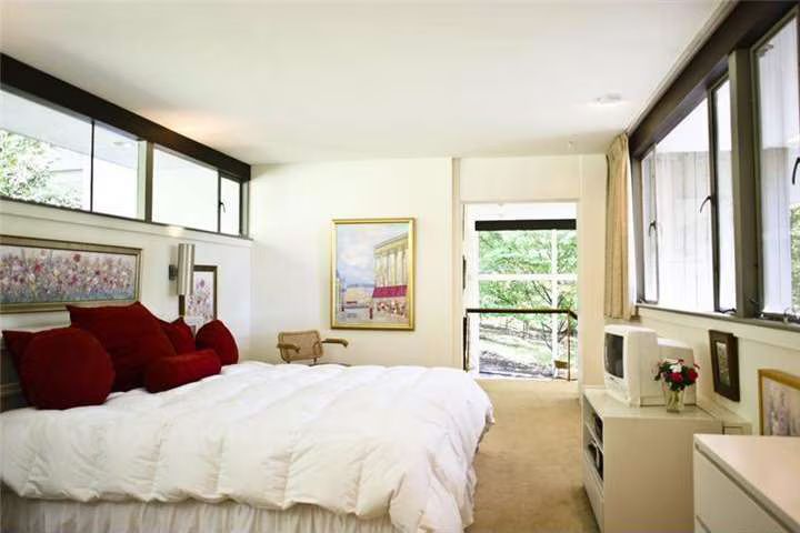 Bedroom with a white bed, red pillows, artwork, a small TV, and large windows overlooking a green outdoor area.