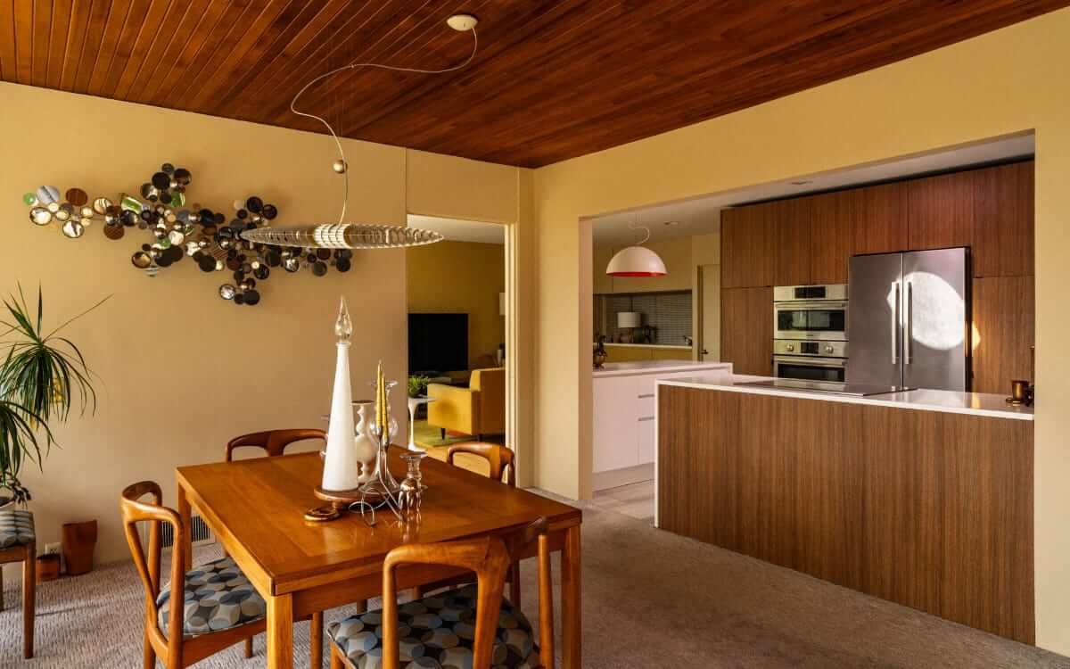 Dining room with wooden table, chairs, and kitchen view. Features a wood ceiling and a modern wall sculpture.