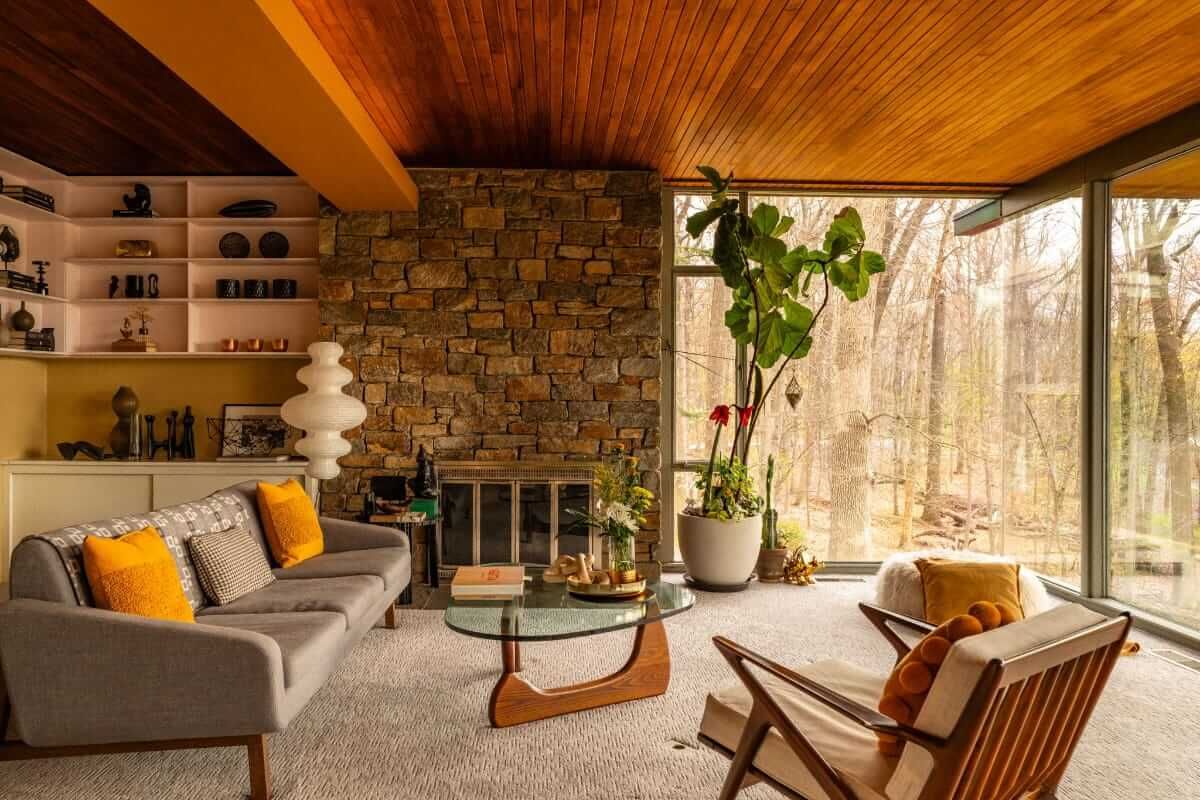 Living room with stone fireplace, wood ceiling, large window with trees, gray couch with yellow pillows.