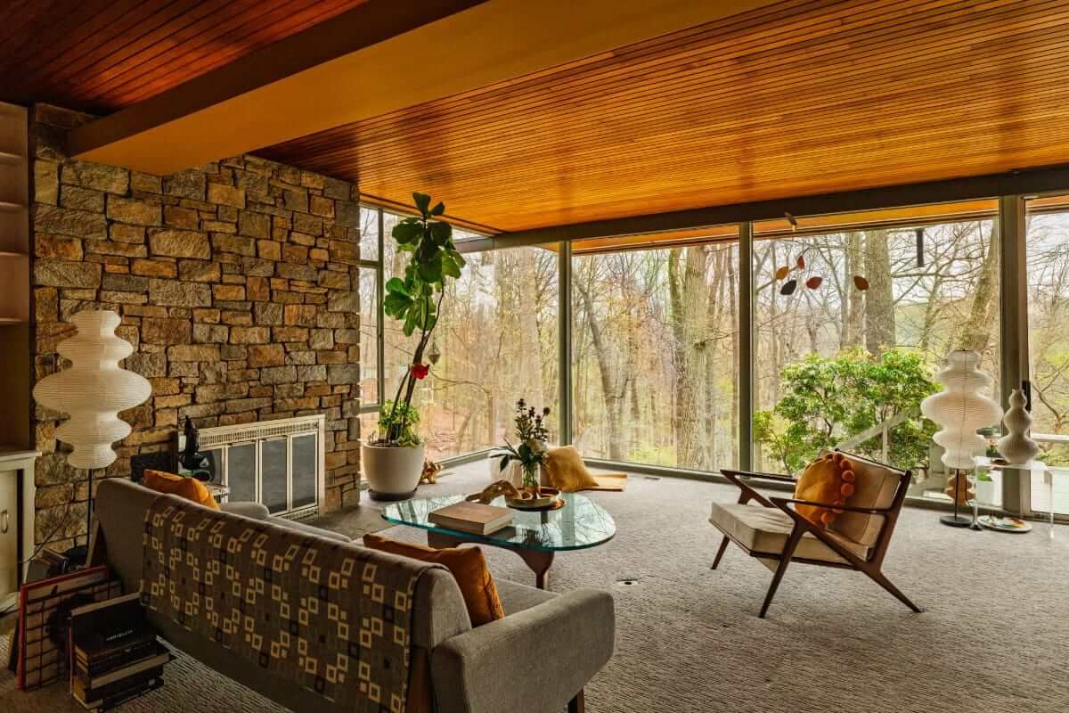 Living room with stone wall, floor-to-ceiling windows, mid-century furniture, and forest view.