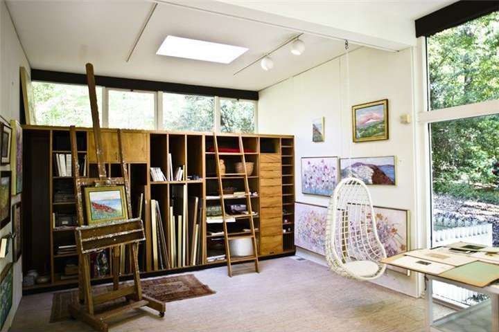 Art studio with easel, storage, paintings, hanging chair, and large windows.