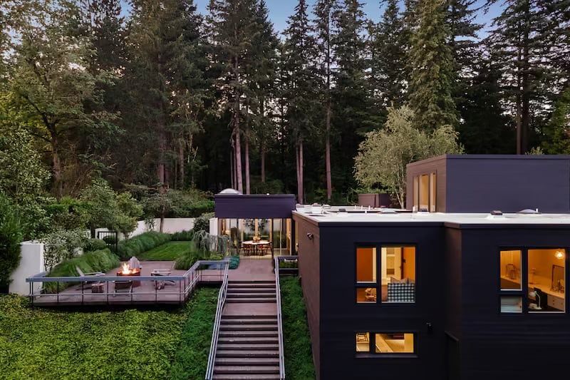 Modern house with dark exterior, wood deck, stairs, fire pit, and surrounding greenery.