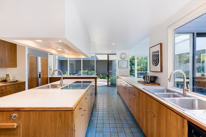 Long, modern kitchen with wooden cabinets, blue tiled floor, and views of a patio through large windows.