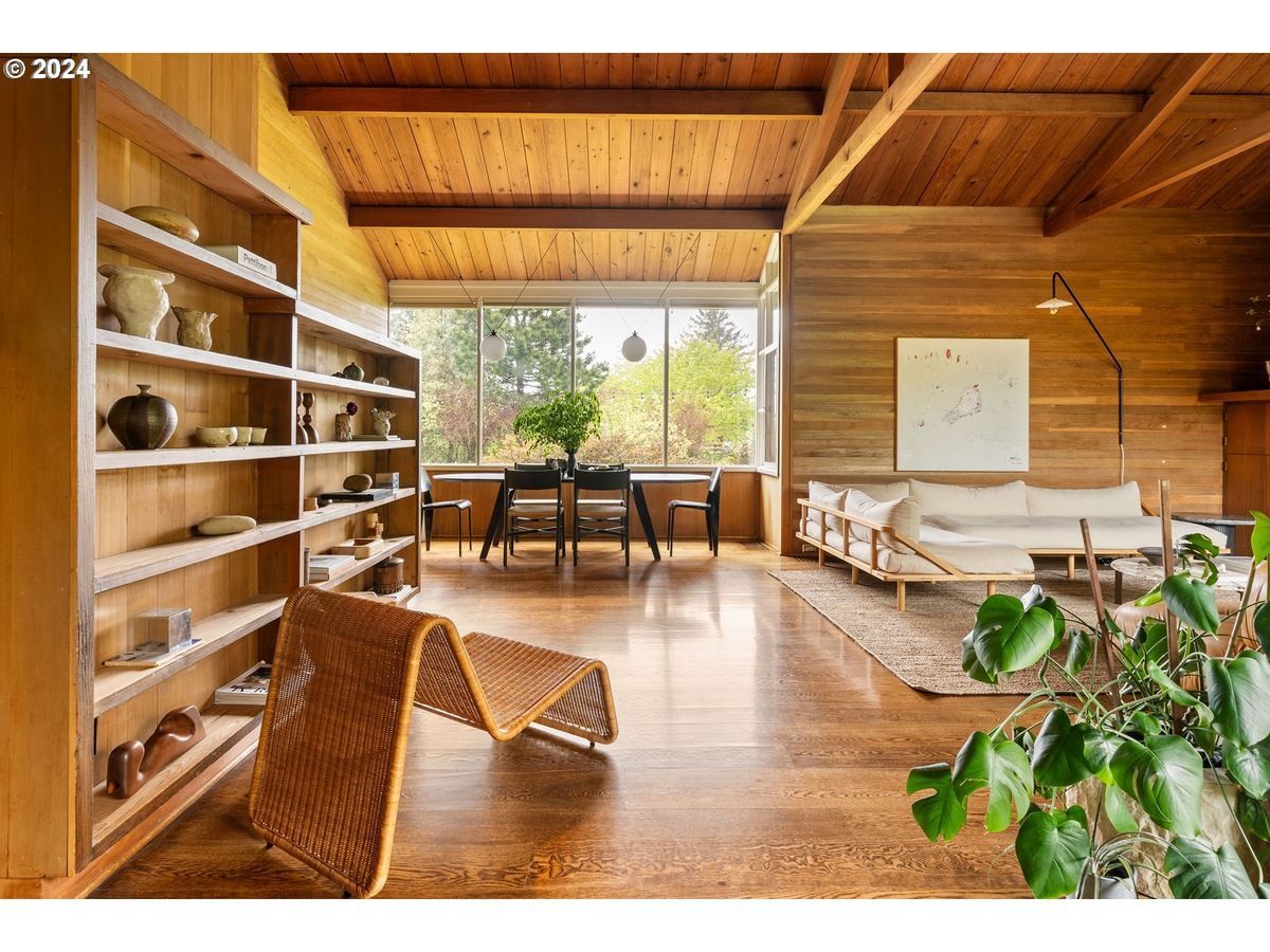 Wooden living room with built-in bookshelves, dining area, and white sofa, with large windows overlooking trees.