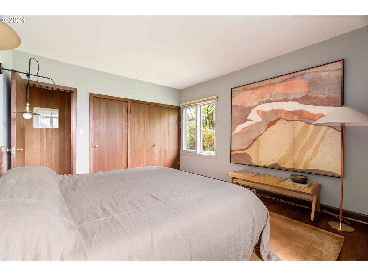 Bedroom with bed, art, wood doors, window, and copper floor lamp. Light gray walls.