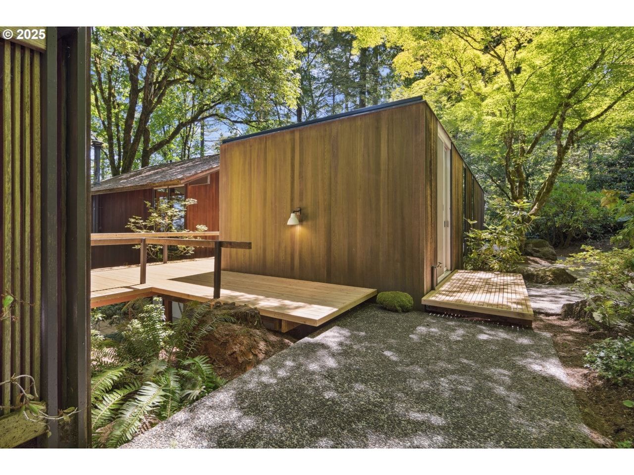 Wooden cabin with deck surrounded by lush greenery and gravel pathway.