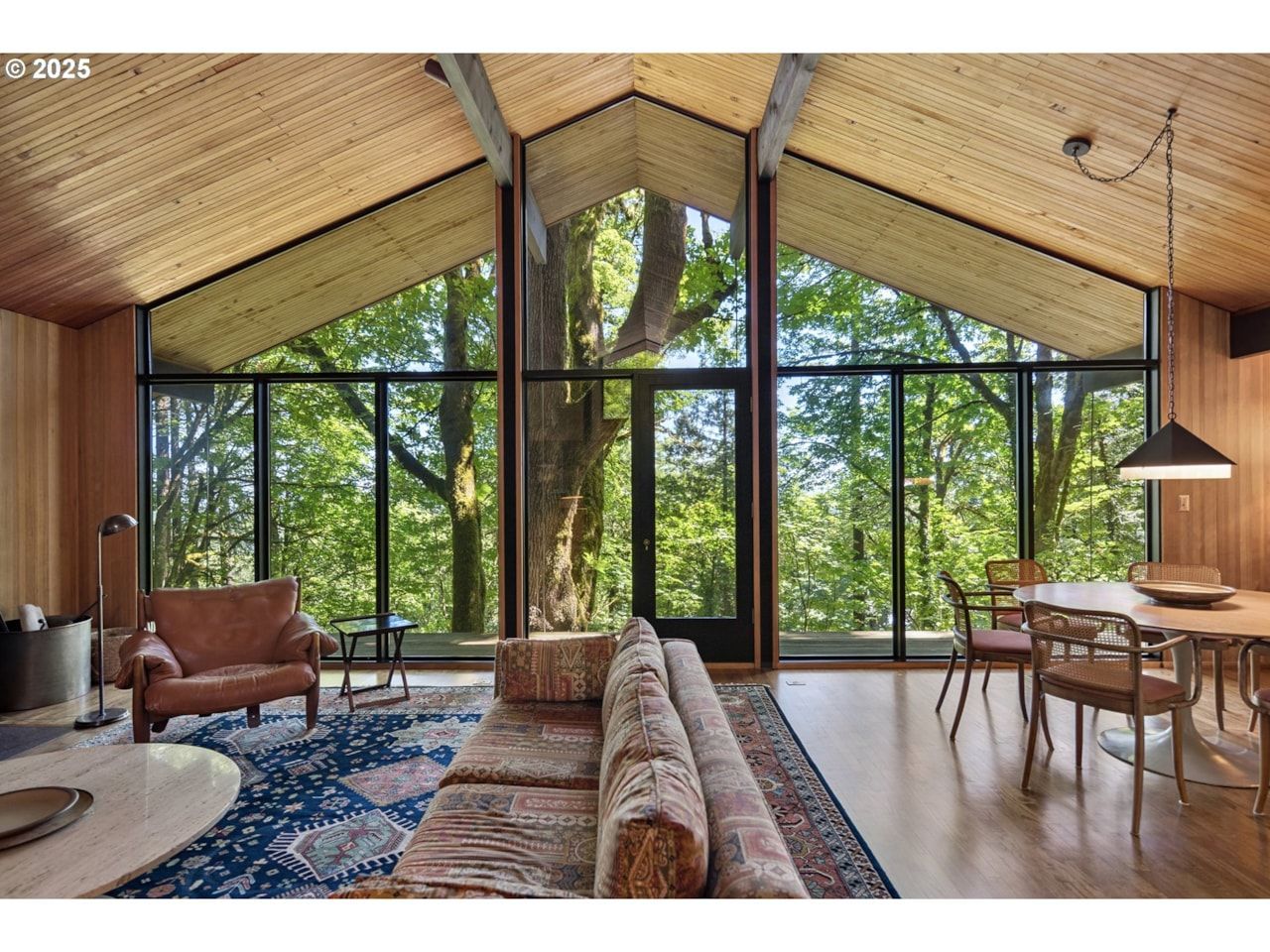 Mid-Century living room with floor-to-ceiling windows overlooking a forest. Wooden ceilings, furniture and accents.