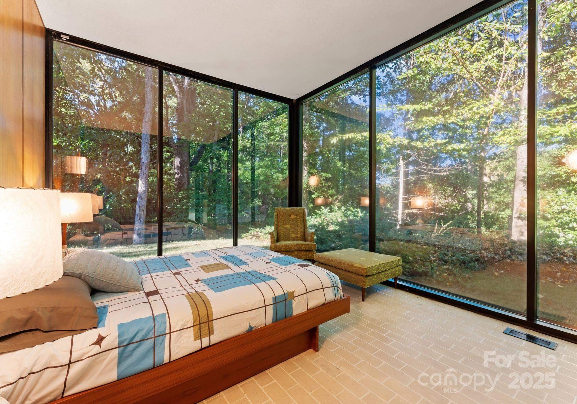 Bedroom with floor-to-ceiling windows overlooking a wooded area. Bed, chair, and ottoman are visible.