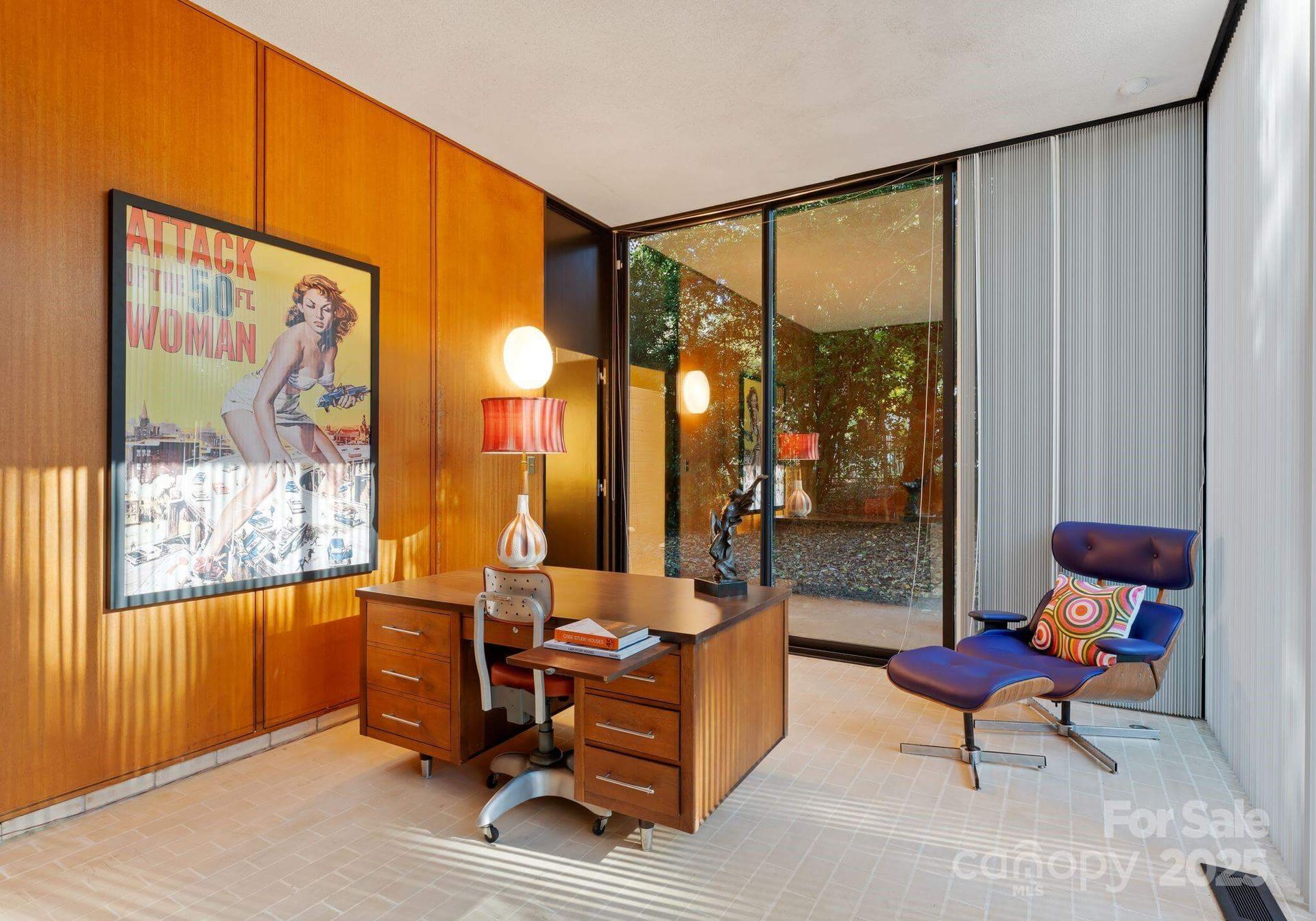 Mid-century modern home office with wooden paneling, desk, and poster; overlooks an outdoor area.