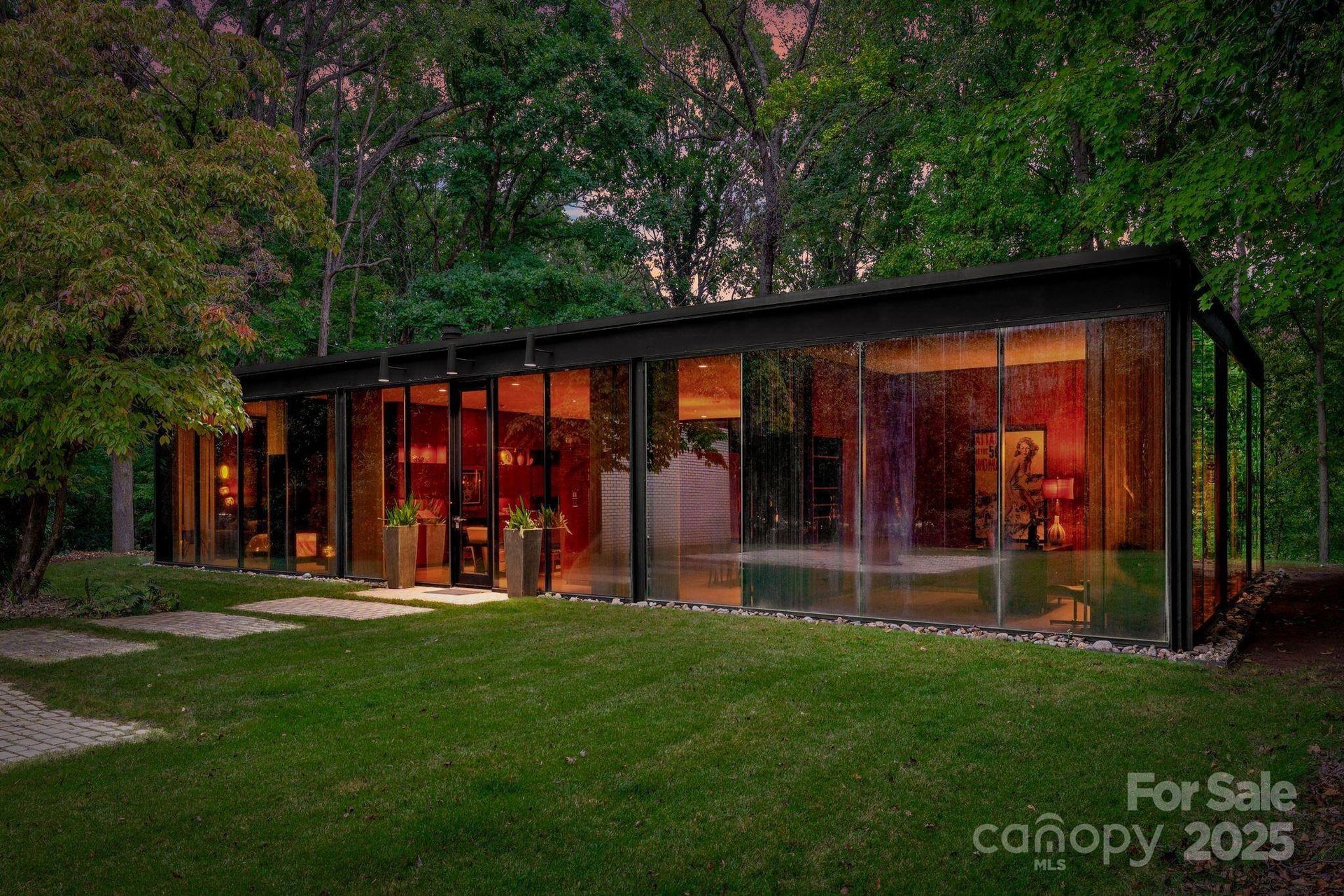 Modern glass house with interior lights reflecting on the transparent walls; surrounded by lush greenery.