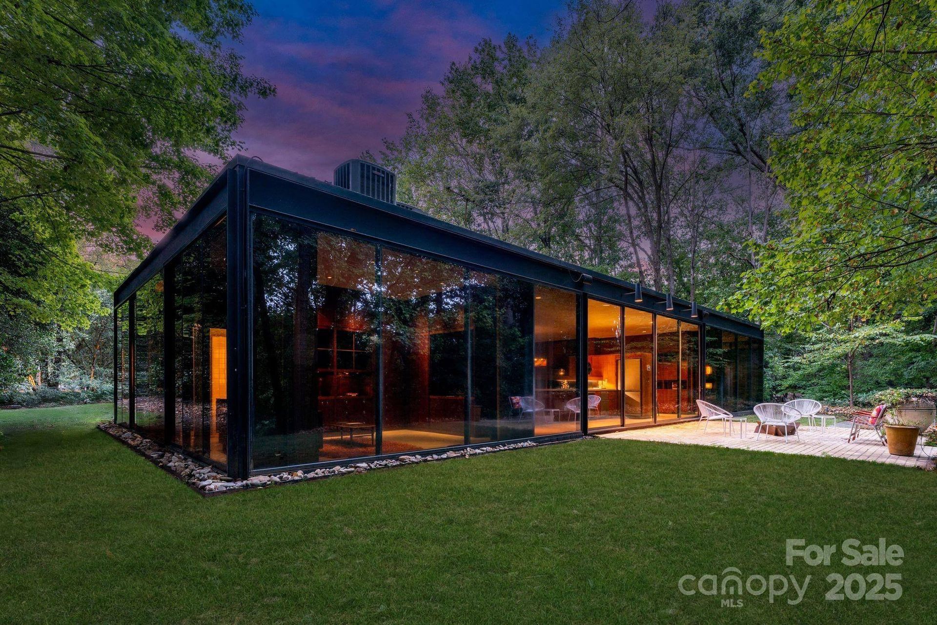 Glass-walled modern home at dusk, on green lawn with trees, 