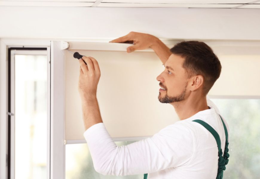 Man installing a roller shade in a window frame, indoors. He wears a white shirt, green straps.