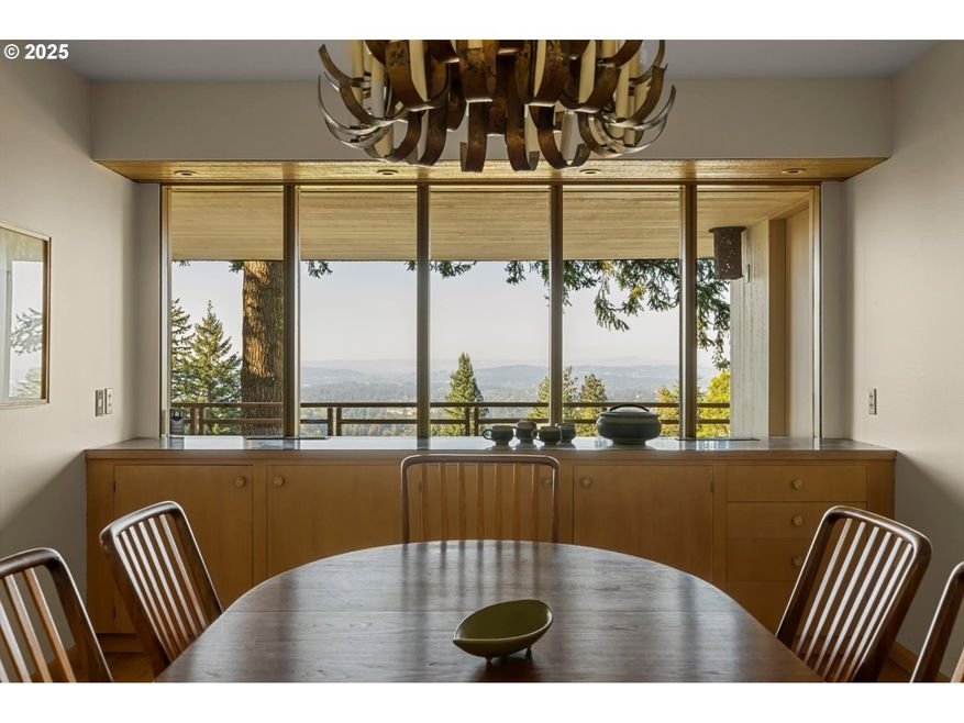 Dining room with large window showcasing a view, wooden table and chairs, and a decorative chandelier.