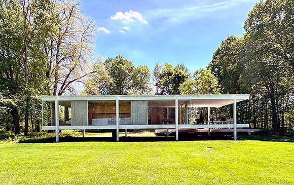 Modern glass and steel house on a grassy lawn with trees in the background under a blue sky.
