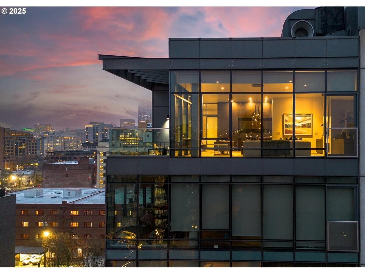 Modern glass building with illuminated interior at dusk, city skyline in the background.