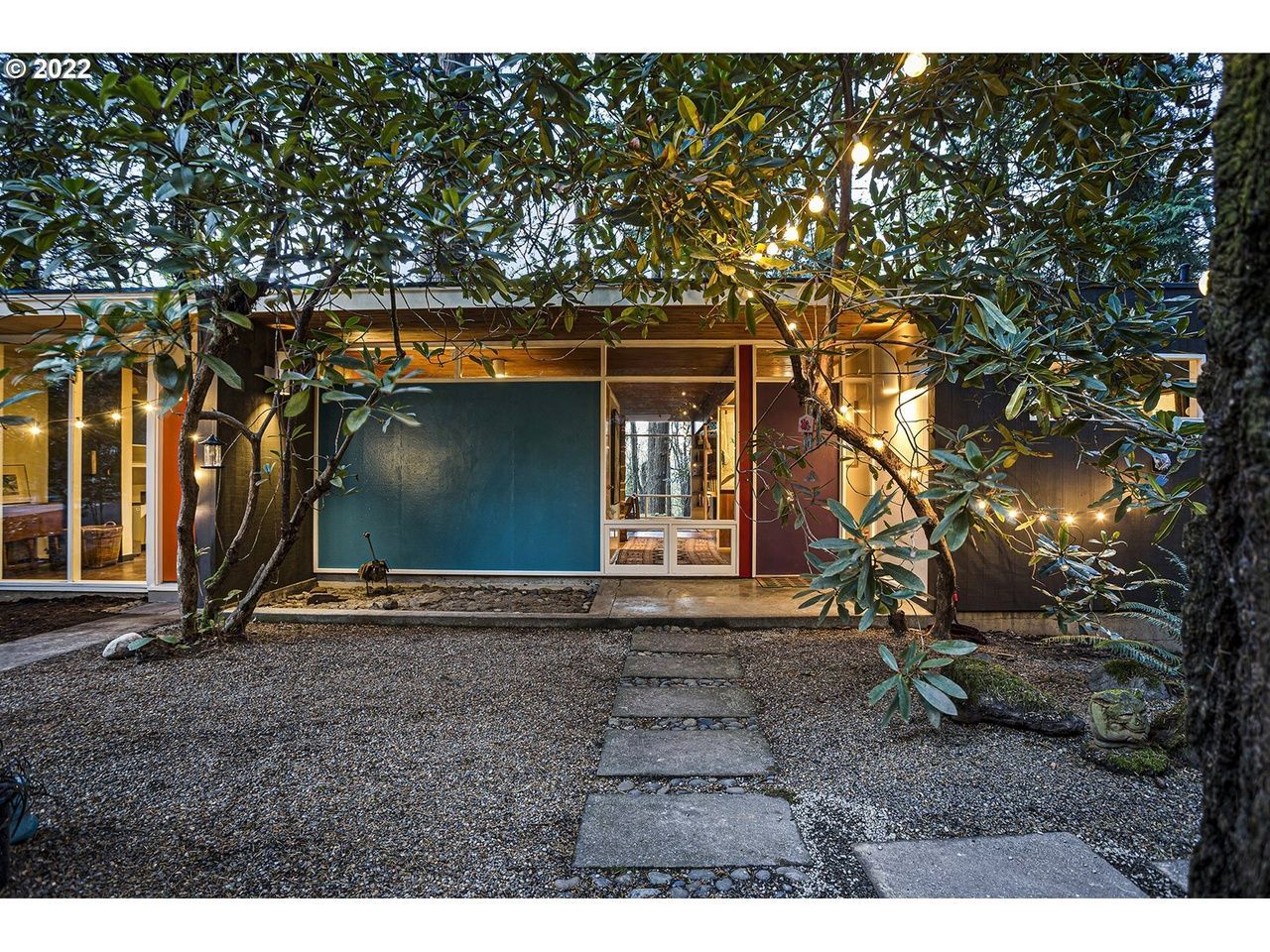 Modern home exterior with gravel yard and stepping stones. Blue, red, and glass walls with string lights in trees.