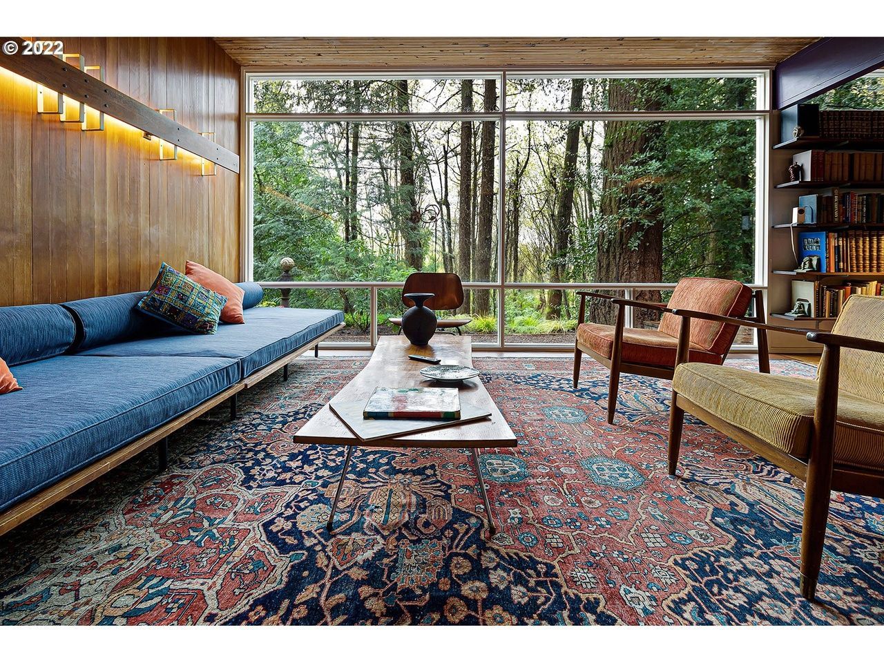 Living room with a large window overlooking a forest; blue couch, patterned rug, armchairs, and wooden paneling.