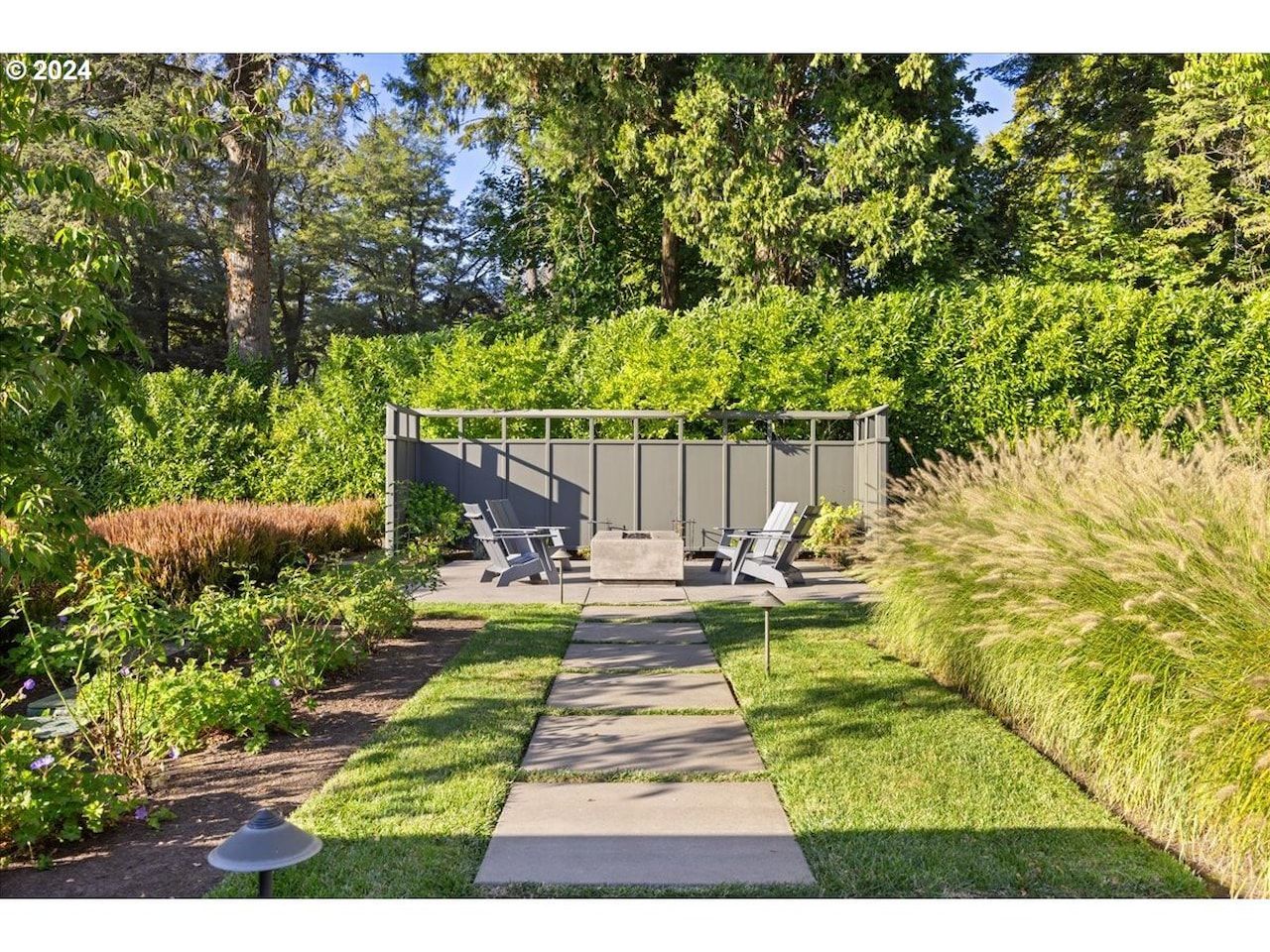 Outdoor seating area with a concrete path, a fire pit, and two chairs surrounded by greenery.