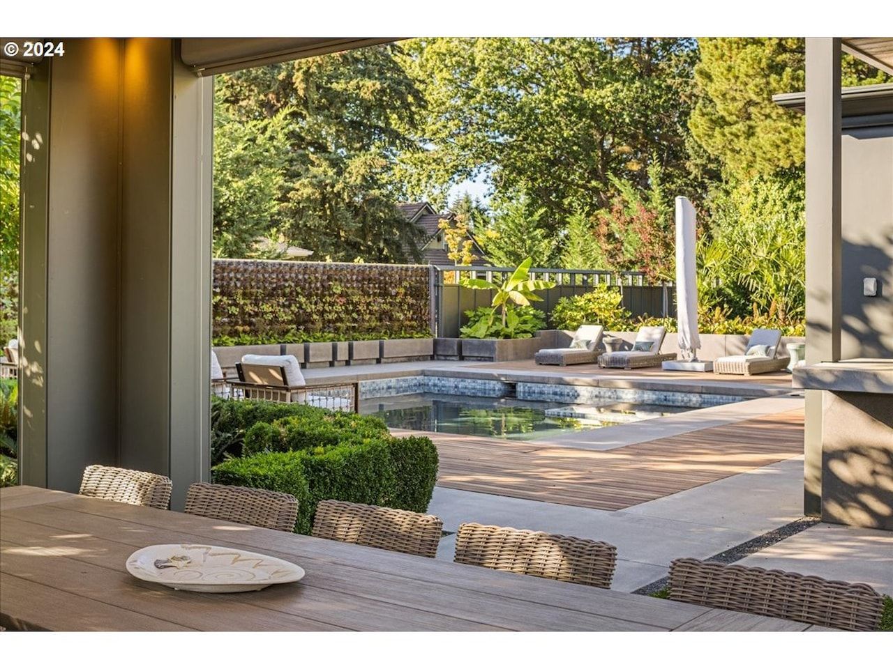 Outdoor patio with pool. Tables and chairs on patio, greenery, and trees in the background.