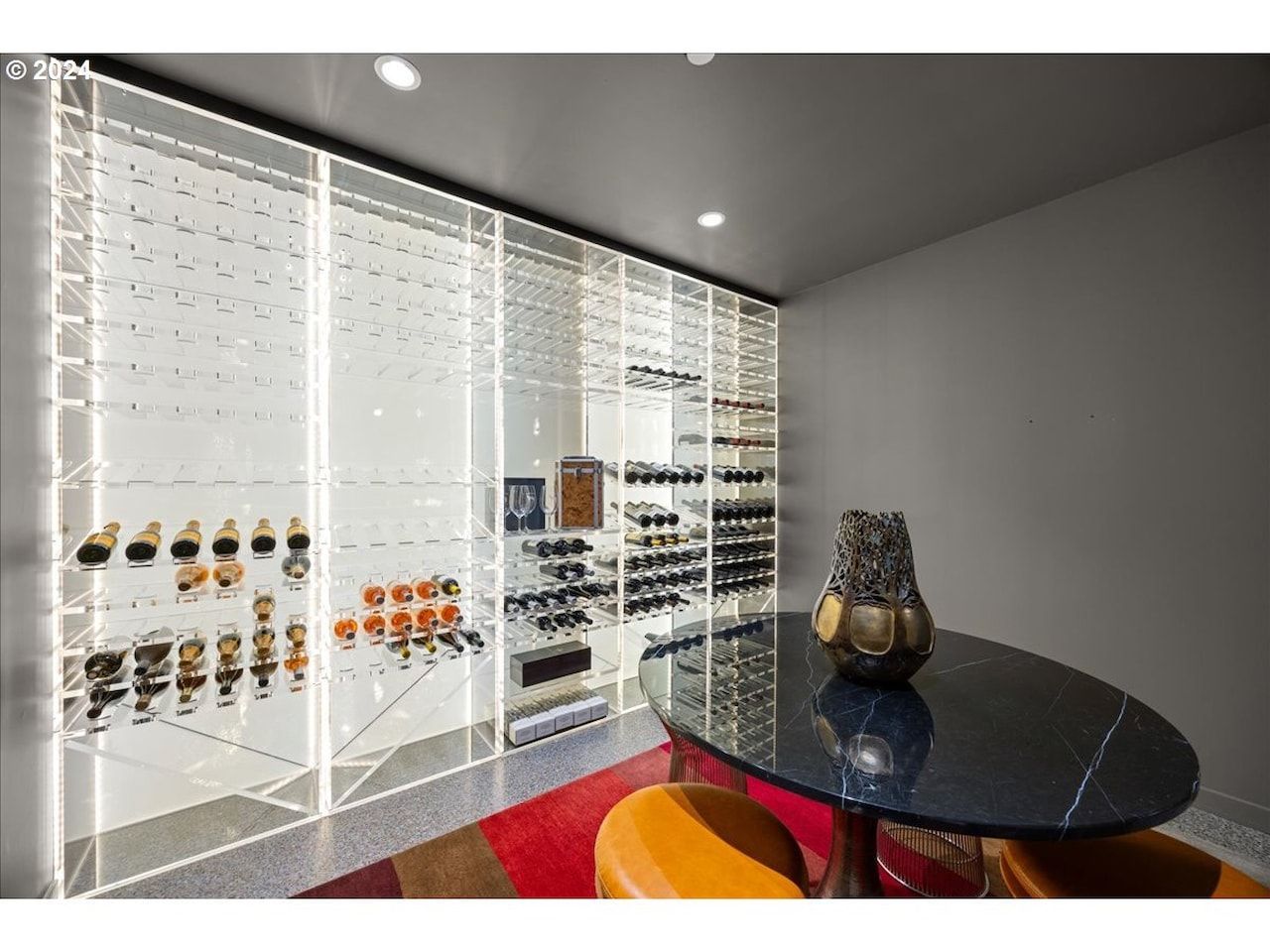 Modern wine cellar with illuminated glass shelving, various bottles, and a table with seating.