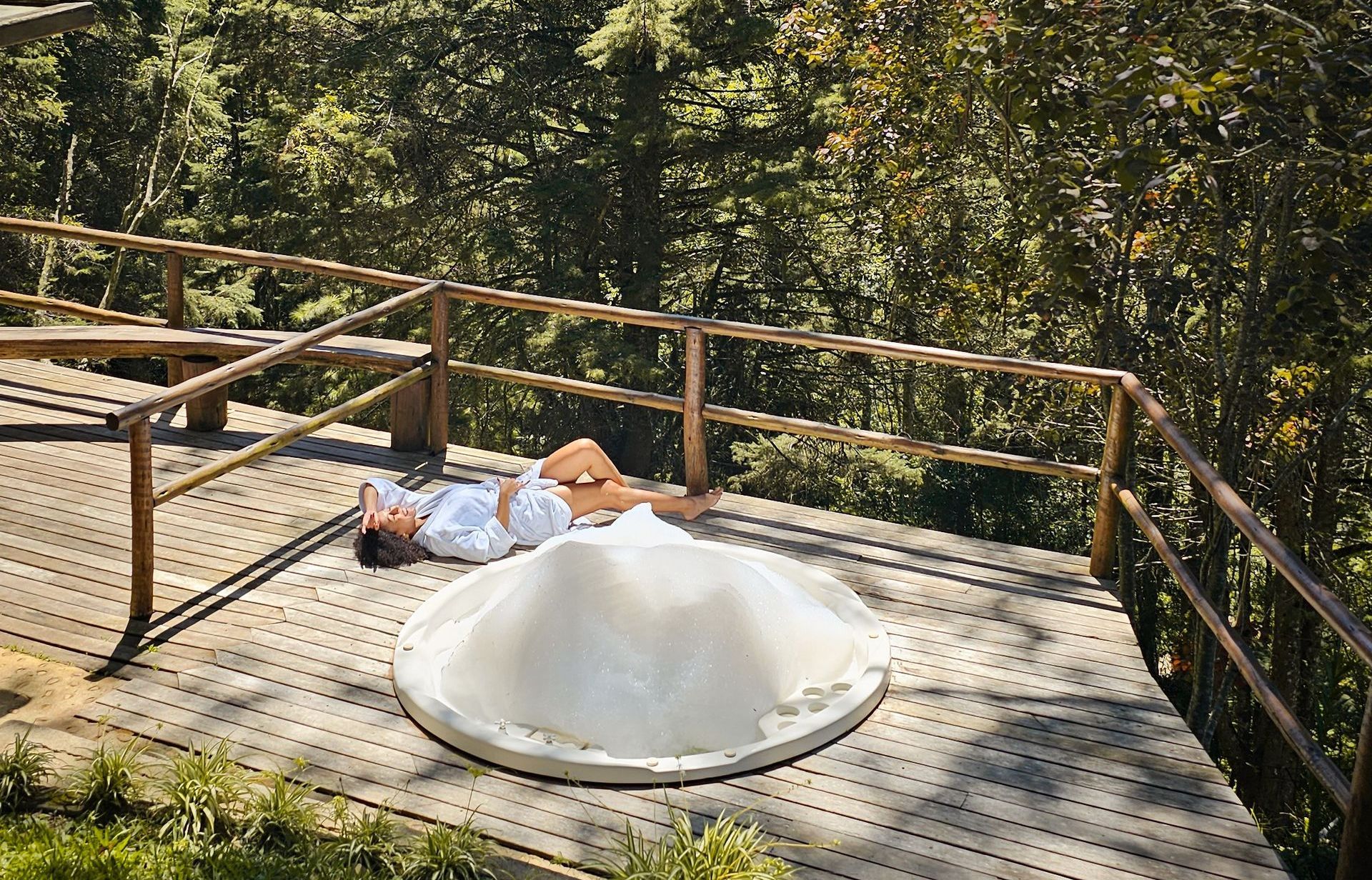 Uma mulher está deitada em um deck de madeira ao lado de uma jacuzzi.
