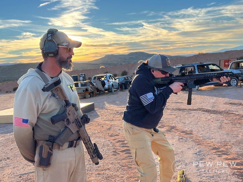 Man with rifle watches another aiming gun at a shooting range in the desert, cars in the background.
