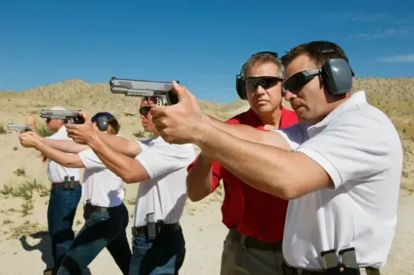 Group of people at a firing range, aiming handguns; all wearing ear protection and sunglasses.