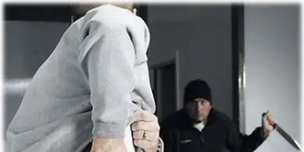 Man in grey hoodie facing another man holding knife indoors.