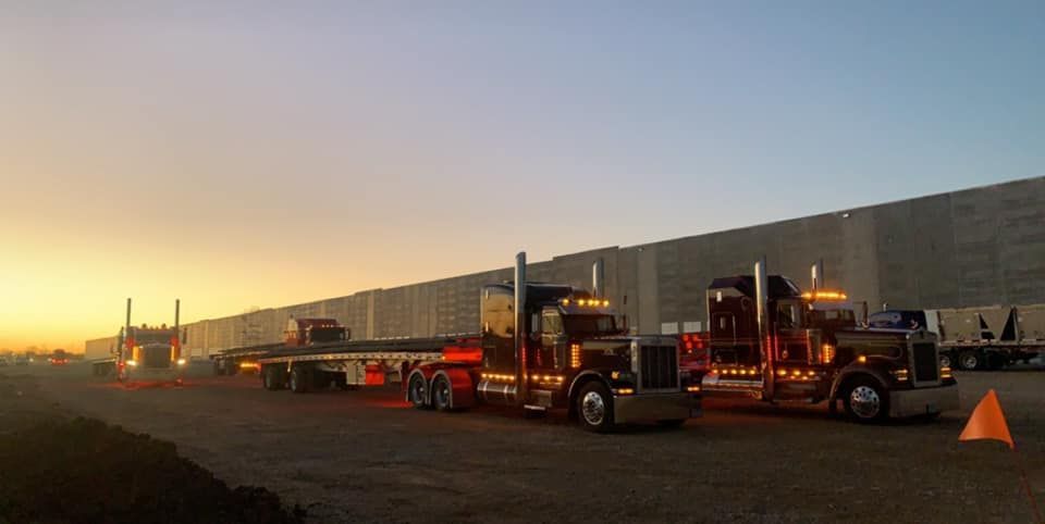 Trucks lined up on a dirt road at dusk with a long building in the background. Orange and yellow colors are present in the sky.