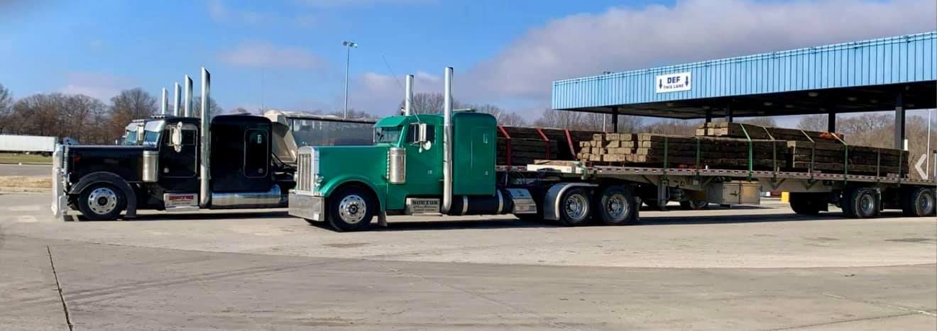 Two semi-trucks parked; one black, one green loaded with lumber, under a covered structure.