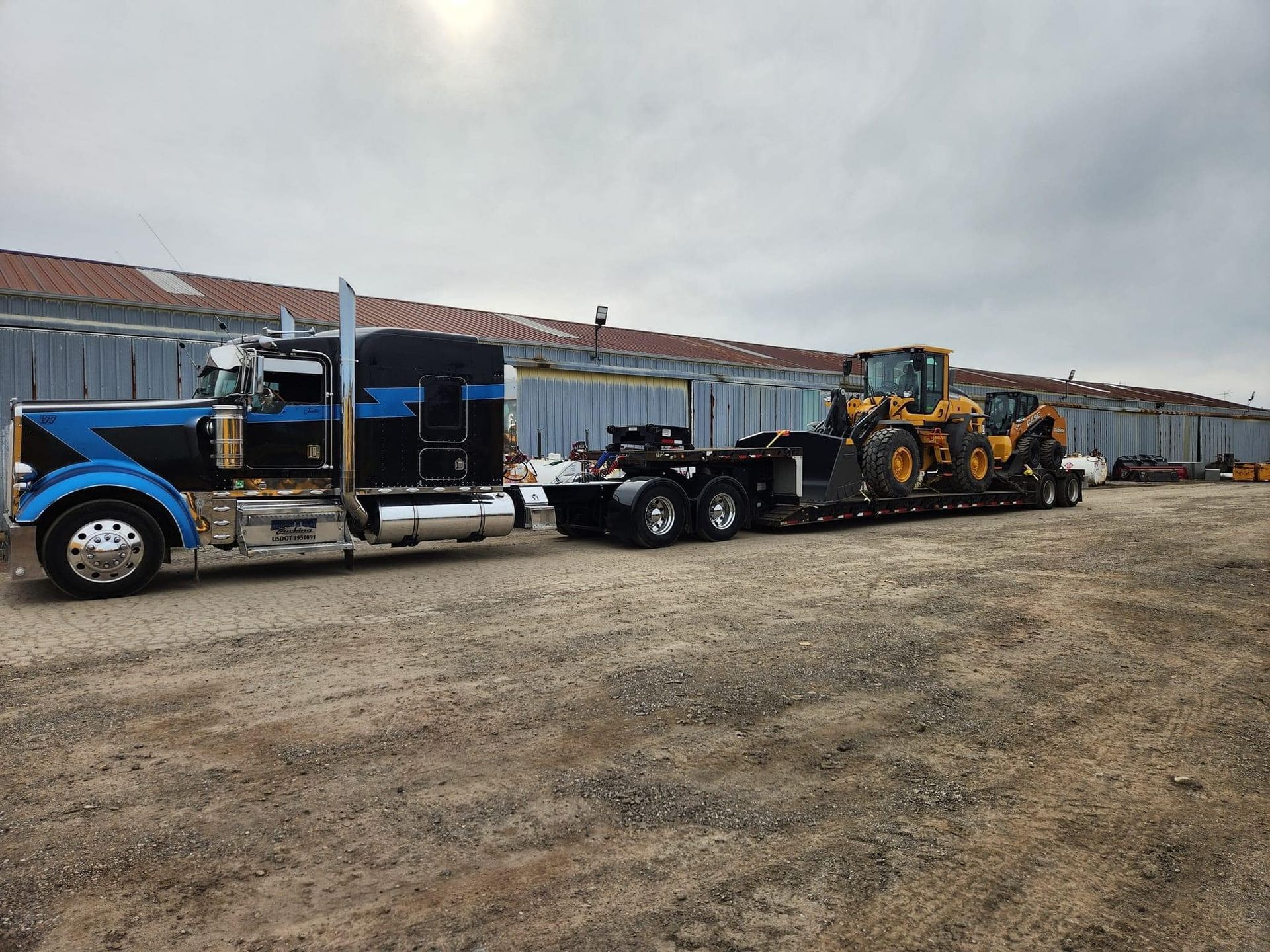 A black and blue semi-truck towing a lowboy trailer loaded with two yellow construction vehicles on a gravel lot.