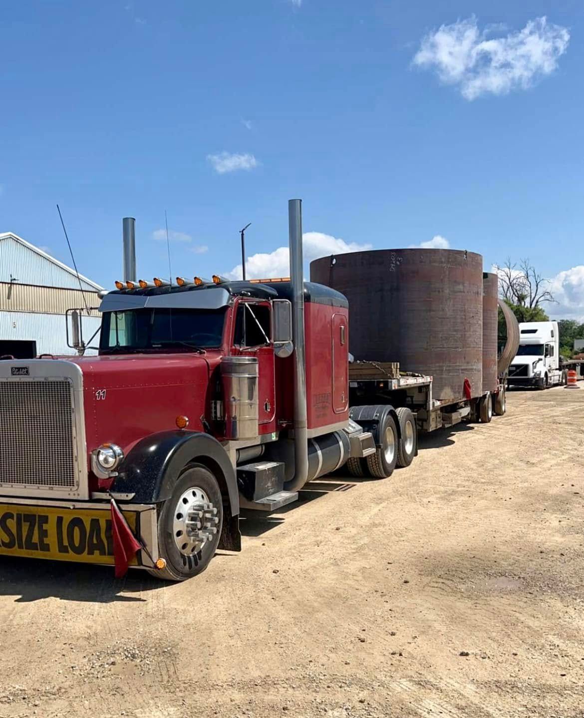 Red semi-truck hauling three large, cylindrical metal tanks on a flatbed trailer.  