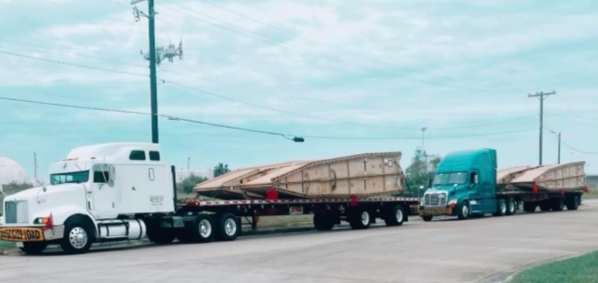 Two semi-trucks hauling large wooden structures on flatbed trailers on a road under a cloudy sky.