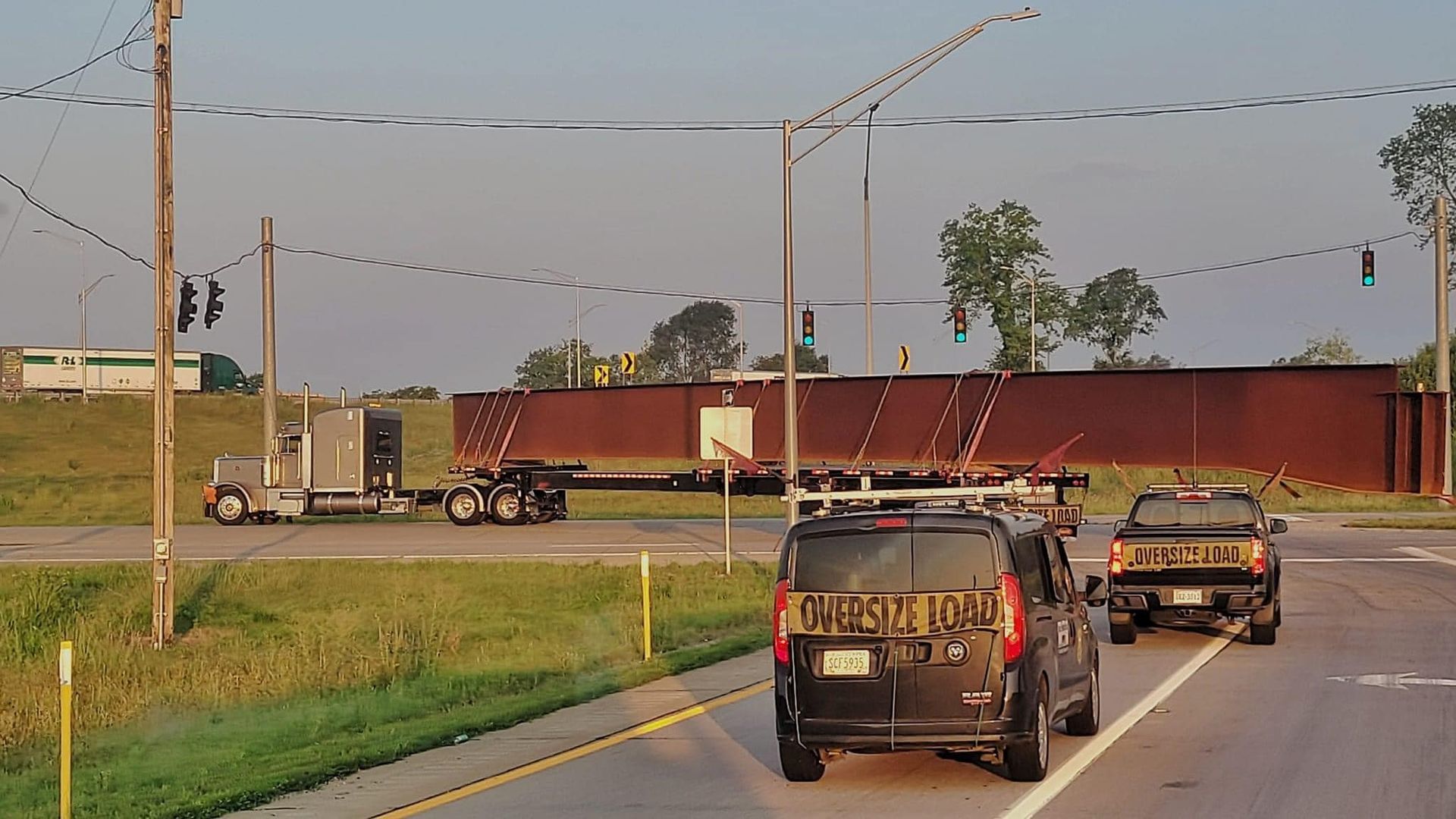 A semi-truck hauls a long steel beam on a flatbed trailer through a roadway intersection. Other vehicles wait at a traffic light.
