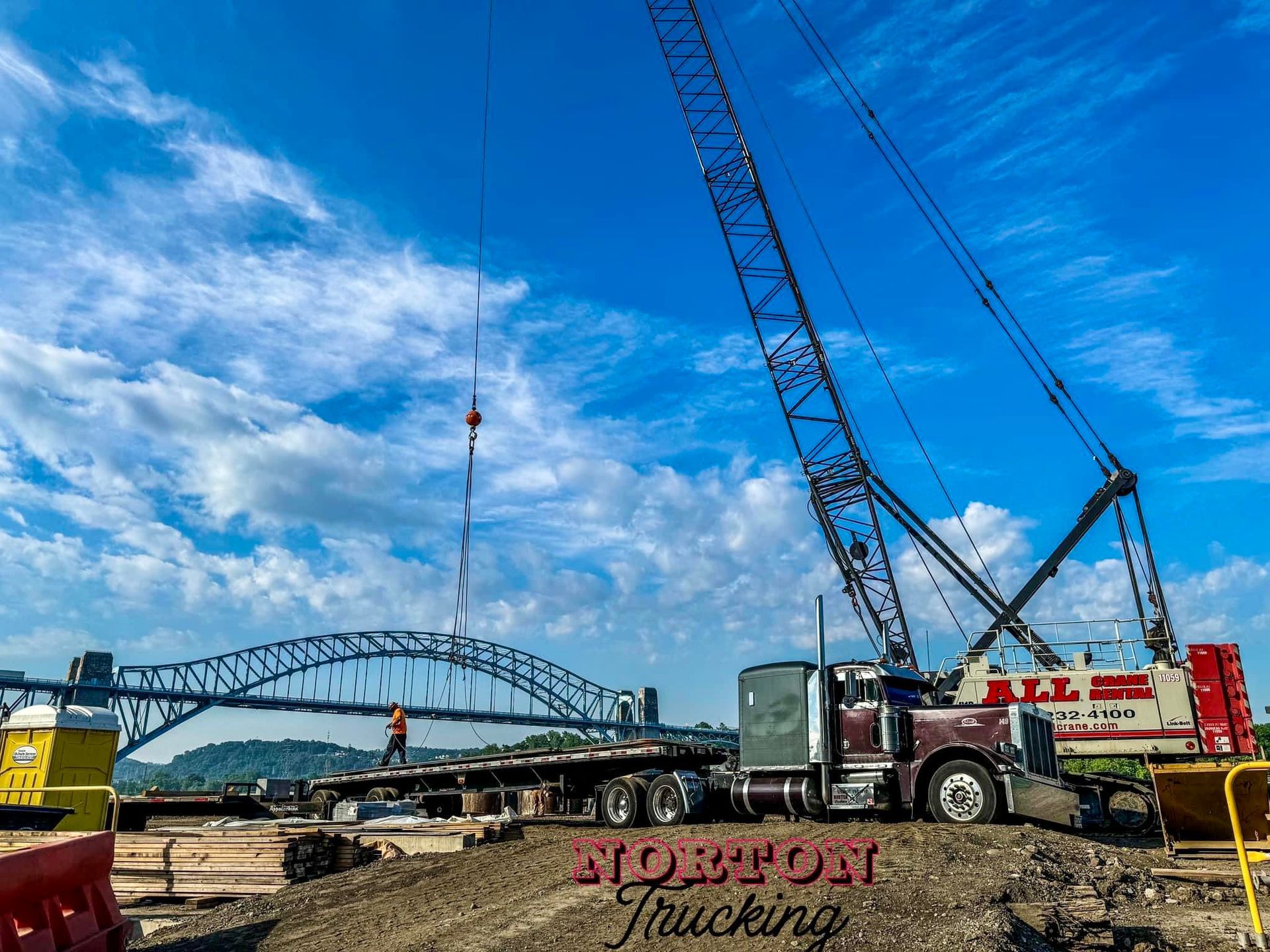 A large crane lifting something onto a flatbed truck at a construction site near a bridge on a sunny day. Norton Trucking logo.