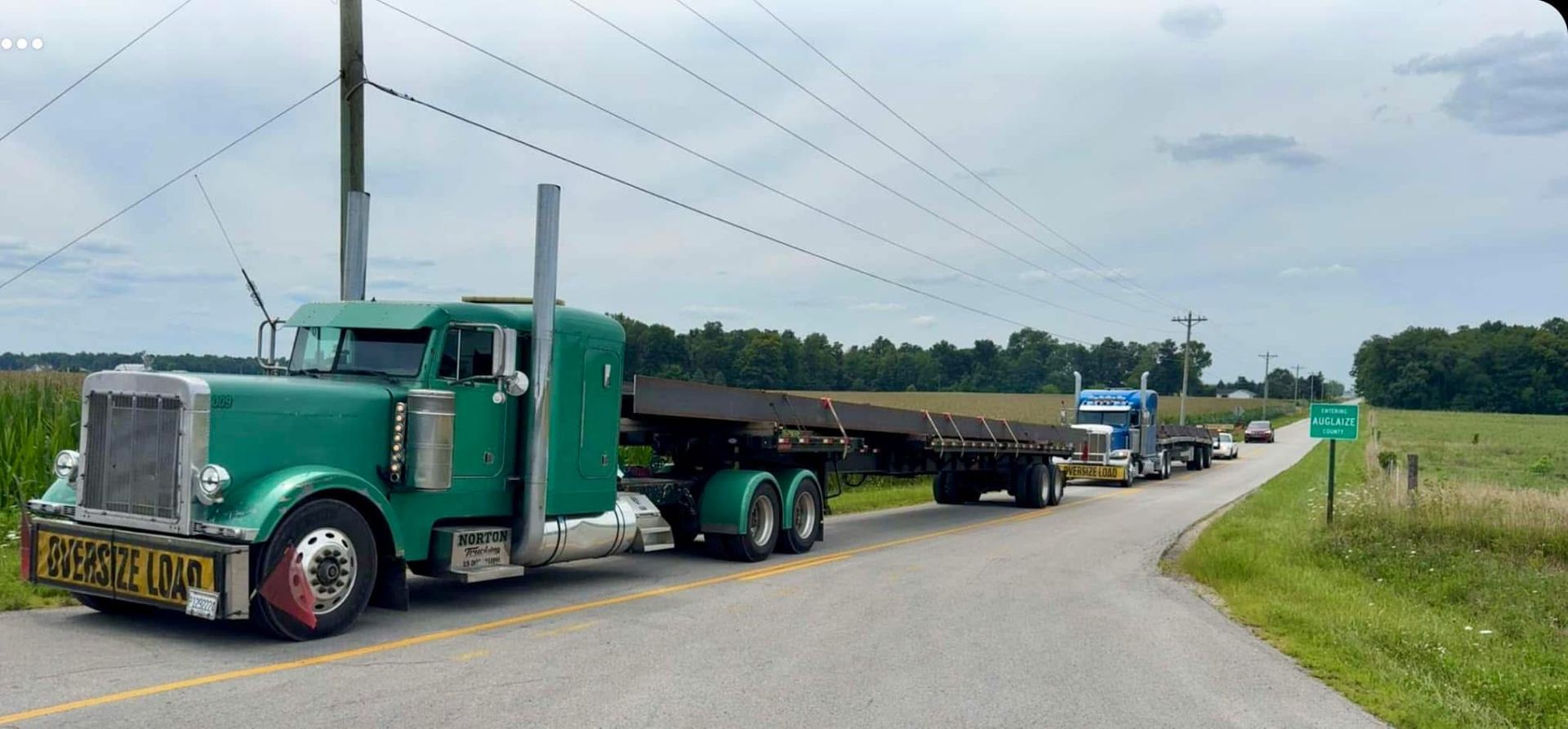 Green semi-truck towing a flatbed trailer on a rural road, followed by other trucks under power lines and overcast sky.