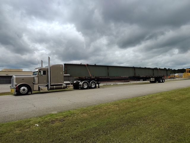 Tan semi-truck carrying a long, dark steel beam on a trailer along a road, cloudy sky.