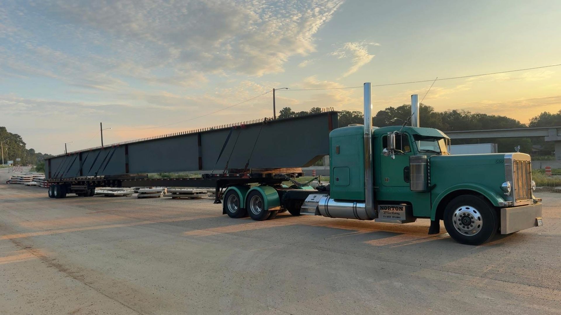 Green semi-truck hauling a long, dark steel beam on a flatbed trailer, likely for construction.