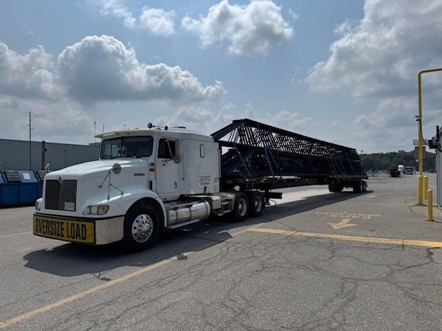 White semi-truck carrying an oversized dark metal structure on a trailer, marked 