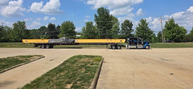 Blue semi-truck hauling long yellow and gray beams on a flatbed trailer across a parking lot.