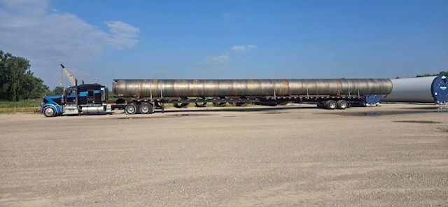 Semi-truck hauling an extra-long cylindrical tank on a flatbed trailer under a blue sky.