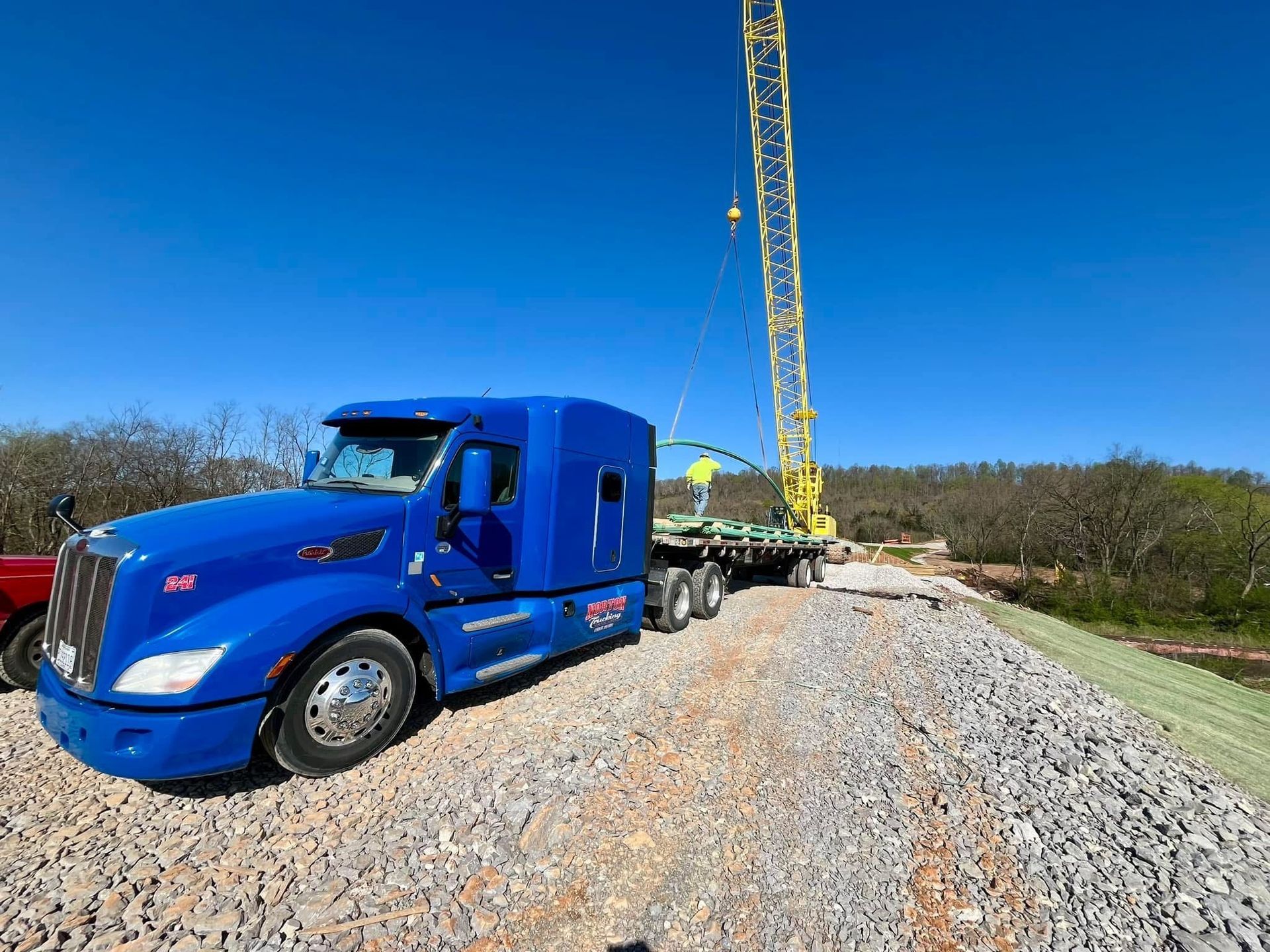 Blue semi-truck and crane unloading materials on a gravel road. The sky is clear and blue.