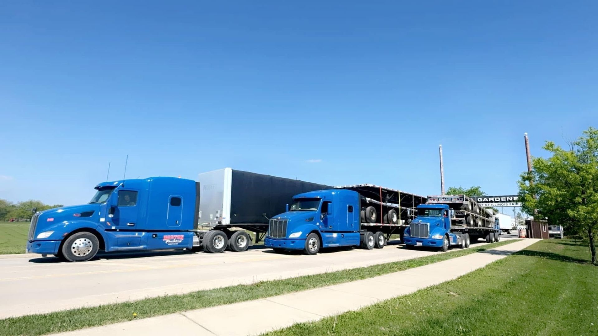 Three blue semi-trucks in a row outdoors on a sunny day, one with a silver trailer and two carrying long loads.